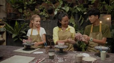 İki farklı gencin el yapımı kil bardakları çömlekçilik atölyesinde masa başı öğretmenine gösterirken geniş açıdan çekilmiş görüntüleri.