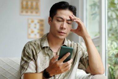 Genç adam kanepede oturuyor ve cep telefonundan mesaj okuyor.