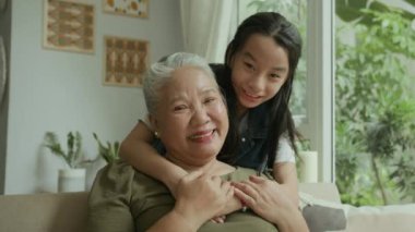 Evde vakit geçirirken kameraya gülümseyen son sınıf öğrencisi ve Asyalı torununun portresi.