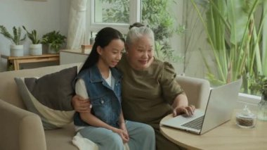 Orta yaşlı ev hanımı ve Asya kökenli torunu oturma odasındaki kablosuz bilgisayarı kullanarak video konuşmaları.