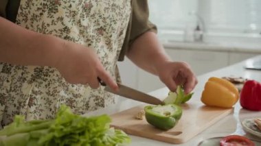 Cropped shot of anonymous housewife in apron cutting bell pepper for lunch at kitchen counter