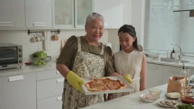 Orta uzunlukta Asyalı neşeli büyükanne ve genç torunun evde taze pizza tutup kameraya gülümsediği bir portre.