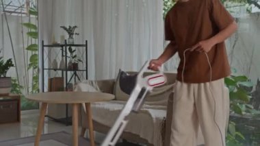 Sıradan giysili Asyalı adamın oturma odasında süpürge temizliği yaparken çekilen görüntüsü.