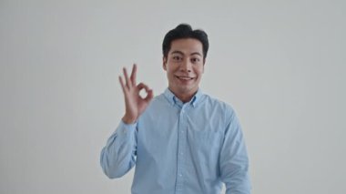 Sıradan mavi tişörtlü Asyalı adamın orta boy portresi tamam işareti ve beyaz arka planda gülümsüyor.