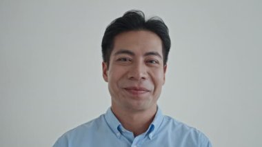 Mavi tişörtlü neşeli Asyalı adamın portresini kapat. Beyaz arka planda kameraya gülümse ve başını salla.