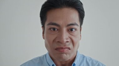 Kapalı çekimde, siyah gözlü ve siyah saçlı Asyalı adam duygularını beyaz arka planda izole bir şekilde kameraya yansıtıyor.