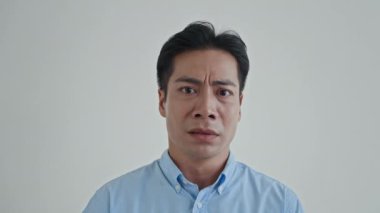 Mavi gömlekli asyalı bir adamın, beyazlar içinde yalnız başına kameraya yakalanmaktan korkmasının yakın plan portresi.