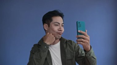 Genç Asyalı adamın mavi arka planda akıllı telefondan görüntüsü.