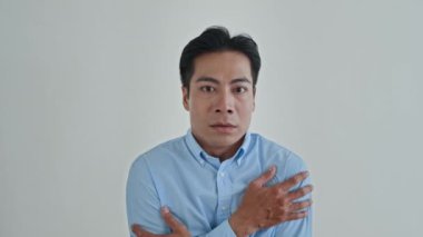 Beyaz arka planda izole edilmiş, korku içinde omuzlarını sallayan mavi gömlekli Asyalı adamın orta boy portresi.