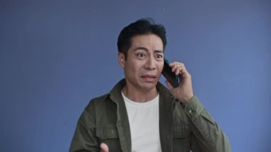 Mavi arka planda izole edilmiş cep telefonuyla konuşurken Asyalı adamın rahatsız olduğu orta ölçekli bir fotoğraf.