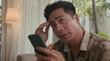 Genç Asyalı adamın evde akıllı telefondan mesaj okurken üzgün olduğu orta boy yakın çekim.
