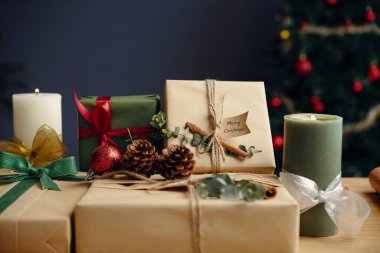 Paketlenmiş hediyeler ve mumlar kozalaklı ve kurdeleli, neşeli bir ortam yaratıyor. Arka planda süsleri olan Noel ağacı
