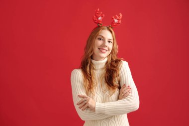 Kırmızı geyik boynuzları ve rahat süveter giymiş, kollarını kavuşturmuş, parlak kırmızı arka planda gülümseyen neşeli tatil havası yaratarak kameraya bakan kadın portresi.