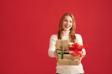 Beyaz kadın kırmızı arka planda duruyor. Gülümsüyor ve çeşitli boyutlarda hediye kutuları taşıyor. Kurdeleler ve dekoratif unsurlarla.