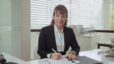 Beyaz ırktan orta yaşlı neşeli bir kadın sosyal hizmet görevlisinin resmi giysi ile ofis masasında doküman doldurma portresi.