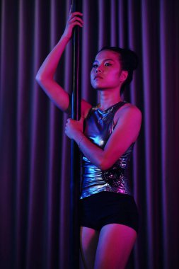 Metalik kıyafetli Asyalı kadın dramatik renkli ışıklandırma altında direk dansı yapıyor, perde arkasına karşı dinamik bir pozda odak ve güç sergiliyor.