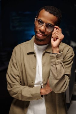 Sıradan kıyafetler giyerken gözlük takan Afro-Amerikan bir adamın portresi. Loş bir ortamda duran, düşünceli bir ifade takınan ve elinde gözlük tutan bir adam.