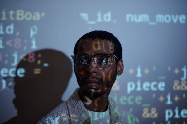 African American man standing against digital code projection on wall, appearing thoughtful and engaged with technology around him