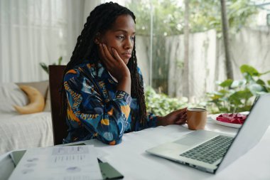 Young woman sitting at laptop, contemplating work with cup of coffee in hand. Bright foliage and large windows create a serene atmosphere for focused working moment clipart