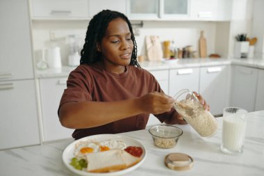 Genç siyahi kadın evde kahvaltı hazırlıyor, mutfak masasına yulaf ekliyor. Parlak mutfak arka planı ve çeşitli kahvaltı malzemeleri.