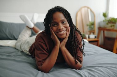 Kahverengi tişörtlü kadın elleri yüzü üzerinde yatarken gülümsüyor. Etrafı ahşap mobilyalar ve ev bitkileriyle çevrili. Sıcak ve misafirperver bir ortam yaratıyor.