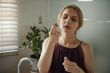 Young woman in bathroom applying makeup, surrounded by potted plants, near window with blinds, wearing a sleeveless top, creating a serene morning routine ambience clipart