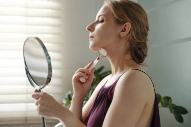 Bir kadın pencerenin önünde yüz silindiri kullanıyor. Aynayı tutuyor, yansımasını düşünüyor. Doğal ışıklandırılmış, sakin bir atmosferi olan bir odada kendine bakımı teşvik ediyor.