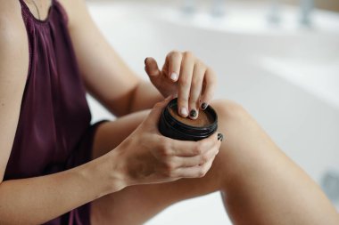 Parlak banyo ortamında koluna krem süren, cilt bakımını gösteren, detaylara dikkat eden, yumuşak odaklı arka planı ve doğal çevresi olan bir kadın.