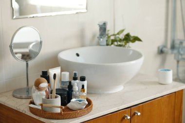 Temiz hatları olan modern banyo kibri ve çeşitli tuvalet eşyaları sepete, yuvarlak aynaya ve zarafete eklenmiş beyaz leğene sahip gösterişli tezgah.