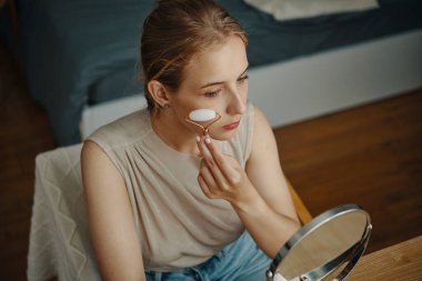 Cilt bakımı için yüz silindiri kullanan beyaz bir kadın. Sandalyeye oturmuş, kendi kendine bakım pratiğine odaklanmış.