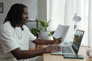 Afrika kökenli Amerikalı bir adam, modern, parlak ofis ortamında dizüstü bilgisayar ve belgeyle çalışıyor. Bitkilerin ve doğal dekorun yer aldığı, üretkenlik sergiliyor.