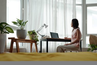 Kadın sakince masasında oturuyor, bitki dolu bir odada, laptopun ve kitapların yanında meditasyon yapıyor, sakin atmosferin tadını çıkarıyor, pencerelerden içeri güneş ışığı giriyor.