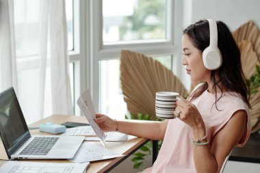 Kadın, dizüstü bilgisayarı açık, kulaklık takmış ve bardak tutarak, doğal elementlerle üretken bir ofis ortamı yaratarak belgeyi inceliyor.