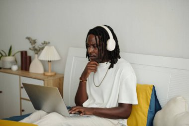 Dizüstü bilgisayarla otururken kulaklık takan kişi elleri çenesinde dalgın dalgın duruyor. Arkaplanda dekor elementleri ve ahşap bir rafta bir lamba var