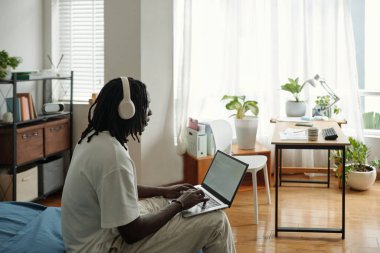 Yatakta kulaklık takan genç bir adam ev ofisinin rahat dizüstü bilgisayarında çalışırken bitki ve ahşap mobilyalarla süslenmiş, odaklanma ve konsantrasyon gösteriyor.