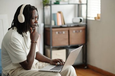 Genç adam evde oturmuş kulaklık takıyor ve dizüstü bilgisayar kullanıyor, ekrana odaklanmış, görünen raf ünitesi ve pencere ışığıyla rahatlamış bir atmosferi var.