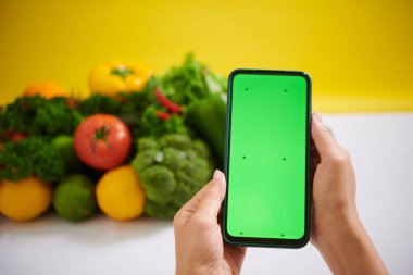 Domates, brokoli ve dolma biber de dahil olmak üzere çeşitli taze sebzelerin önünde yeşil ekran ile el ele tutuşan akıllı telefon.