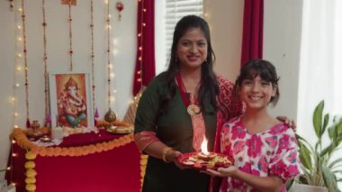 Evde Diwali 'de yanan bir mumla tepsiyi tutarken kameraya gülümseyen neşeli Hintli anne ve kızının portresi.