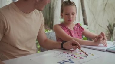 Beyaz bir babanın evde otururken kızına alfabe öğrenmesinde yardım ettiği orta boy bir fotoğraf.