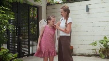 Orta boy beyaz bir anne kızının okula hazırlanmasına yardım ederken, çantasını uzatırken ve açık havada sıcak bir şekilde giyindiğinden emin olurken.