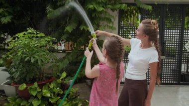 Kafkasyalı bir kızın, annesiyle bahçesinde hortumla bitkileri sularken orta uzunlukta bir pozu.