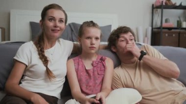 Beyaz kökenli anne babasıyla birlikte evde korku filmleri izleyen orta boy bir kız resmi.