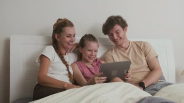 Kafkasyalı ebeveynlerin ve kızlarının aile hafta sonu yatağında dinlenirken dijital tabletle oyun oynamalarının orta uzunluktaki fotoğrafı.