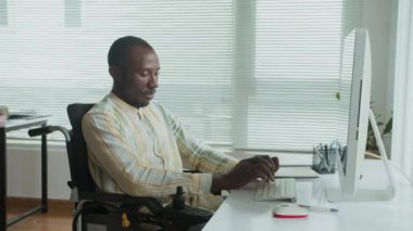 Tekerlekli sandalyede bilgisayar başında çalışan ve ofiste kameraya gülümseyen Afrikalı Amerikalı erkek yöneticinin geniş portresi.