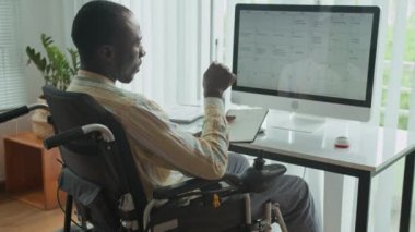 Orta boy Afro-Amerikan iş adamının tekerlekli sandalye kullanarak ofis masasında programını kontrol ederken görüntüsü.
