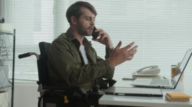 Tekerlekli sandalyedeki beyaz işadamının ofis masasında dizüstü bilgisayarla iletişim kurarken yan görüntüsü.