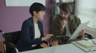 Modern ofiste masa başında otururken iş tartışan iki farklı tekerlekli sandalyeli iş arkadaşının el kamerası görüntüsü.