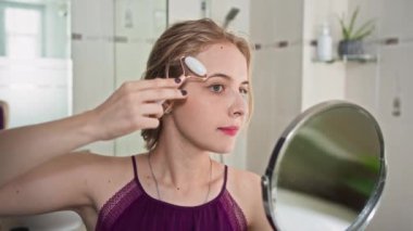 Orta ölçekli, ev elbiseli, aynaya bakarken yüzüne silindirle masaj yapan beyaz bir kadın.