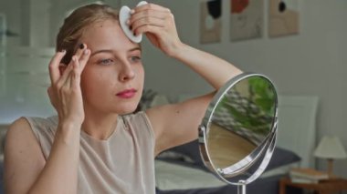 Evinde aynanın önünde yüzüne aletle masaj yapan beyaz bir kadının orta ölçekli fotoğrafı.