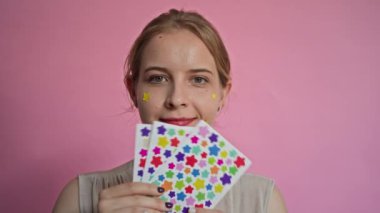 Elinde çıkartmalar olan neşeli kadın zoomcunun orta boy portresi ve pembe üzerine izole edilmiş kameraya gülümsüyor.